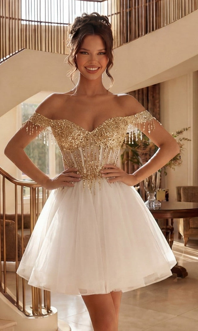 A woman stands indoors, smiling with hands on hips, wearing the Short Champagne Corset Prom Dress: Juliet JT947L in White/Gold, featuring an off-the-shoulder design with a gold sequined bodice and a white tulle skirt.